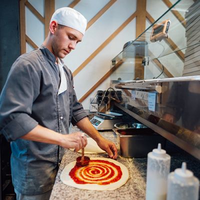 Cook spreads tomato sauce on pizza dough . Catering kitchen work. (Demo)